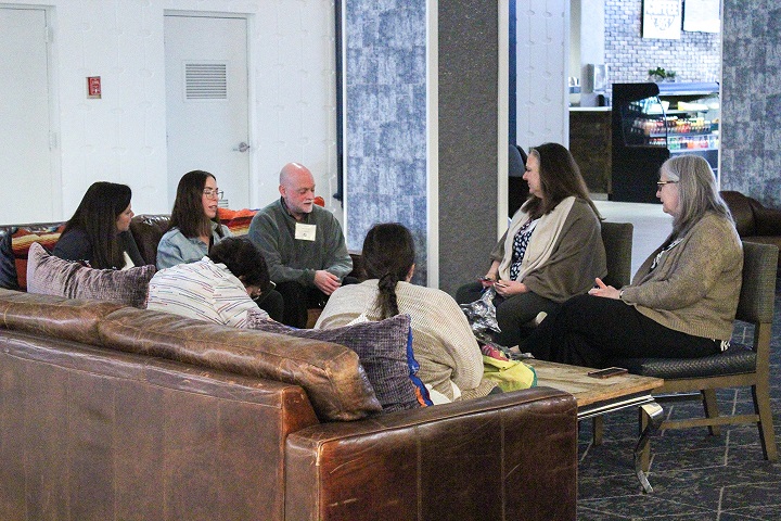 A casual meeting scene in a lounge area. A group of professionals sits on and around a large leather couch, engaged in a focused and relaxed conversation. 
