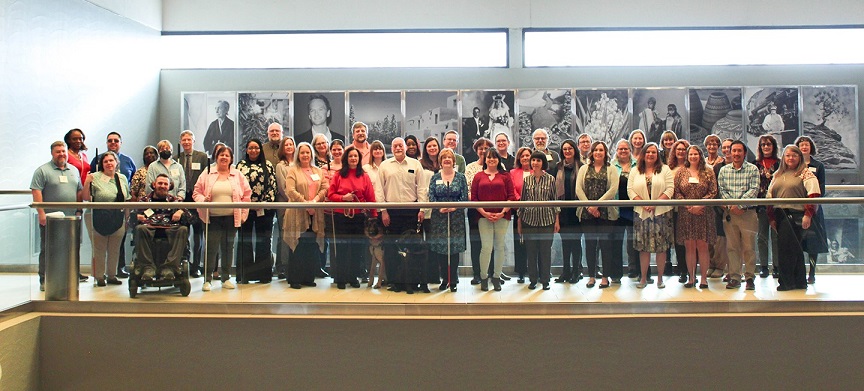 A group photo of conference attendees on a balcony. A diverse group is smiling for the camera, with several black and white framed photographs behind them.